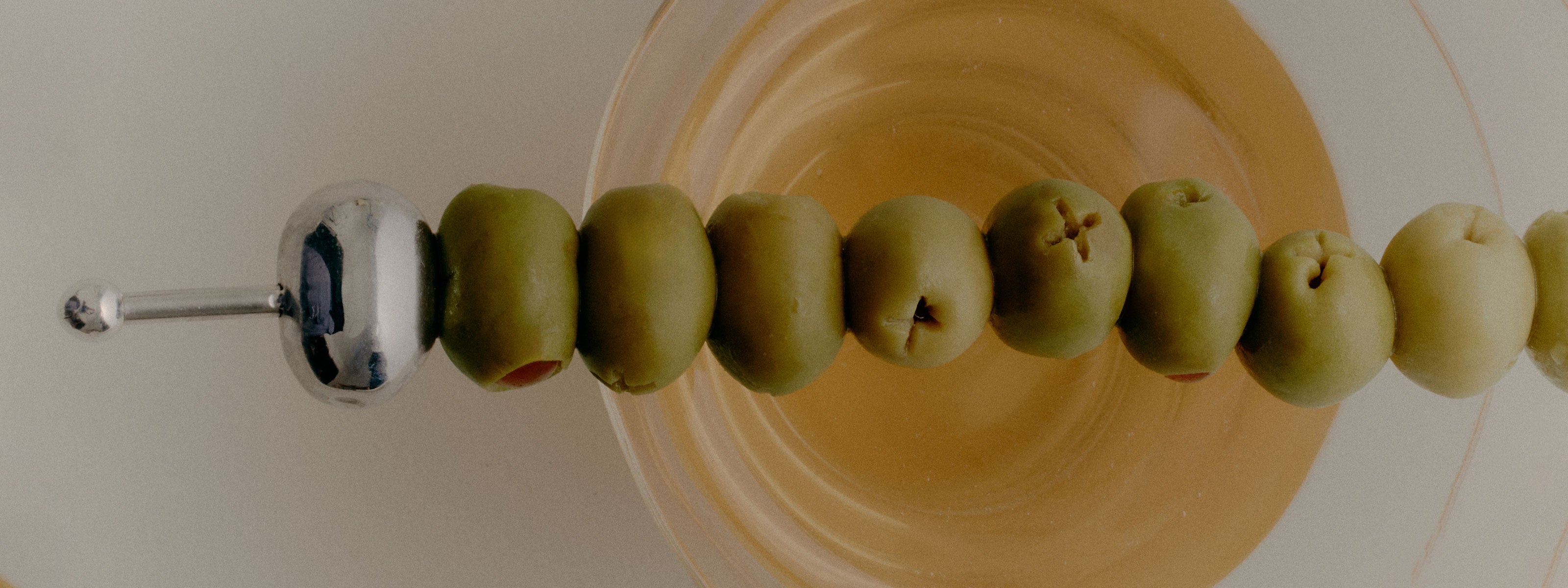 COREY ASHFORD L'aperitif Olive Cocktail Pick in Stainless Steel sitting atop a martini glass with dirty martini and multiple olives