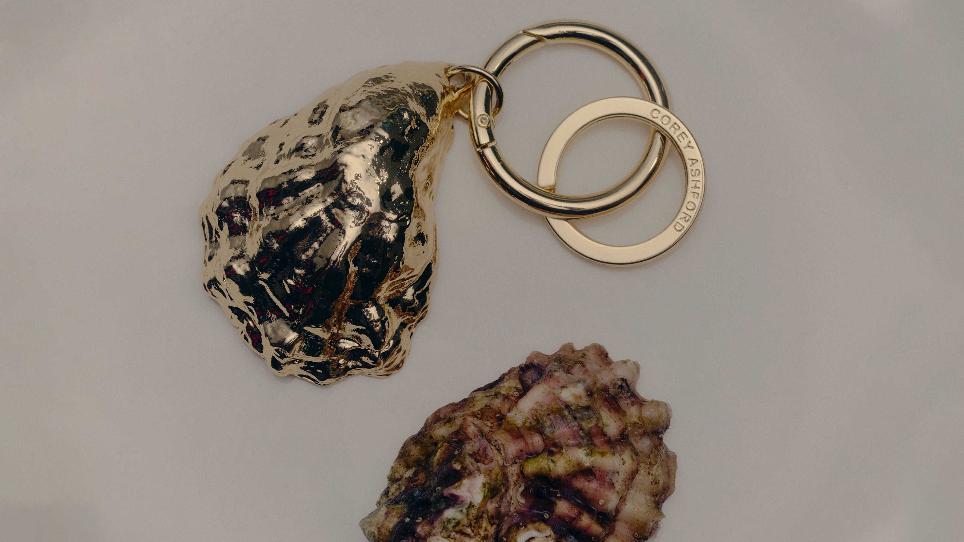COREY ASHFORD L’Apéritif - gold Oyster Keyring with split ring and clasp, photographed flatlay beside a natural oyster shell.