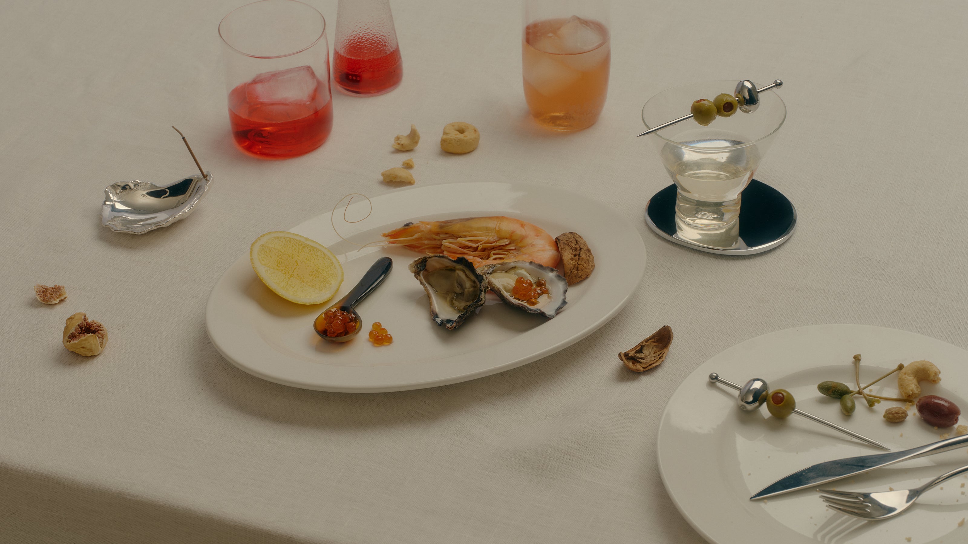 COREY ASHFORD L’Apéritif - oysters and prawn on a white plate with horn caviar spoon, silver oyster incense holder, and a martini glass with a stainless steel olive cocktail pick on a linen table.