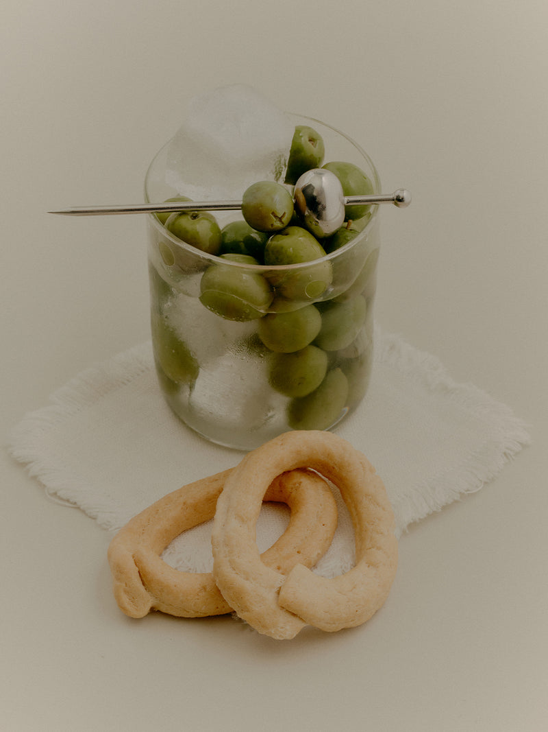 COREY ASHFORD silver Olive Cocktail Pick in stainless steel atop a Malfatti hand-blown Rocks Glass. Modern take on a dirty martini over ice with lots of olives.
